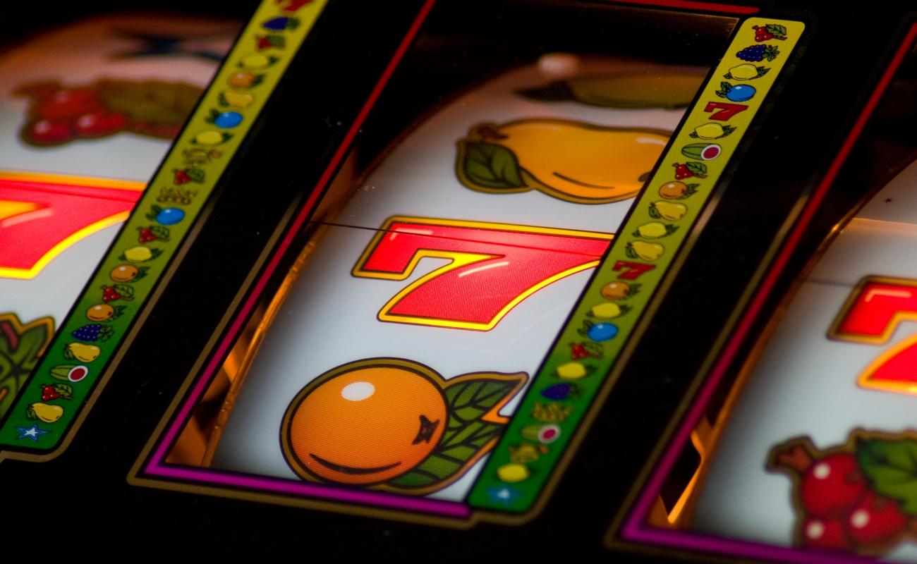 A close-up of the reels on a slot machine. There are three red and yellow sevens across the middle row. Other fruit symbols, such as oranges and cherries, are also visible.