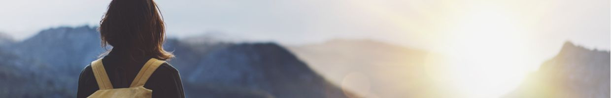 A traveler wearing a backpack overlooks a mountain range with the sunrise in the background.