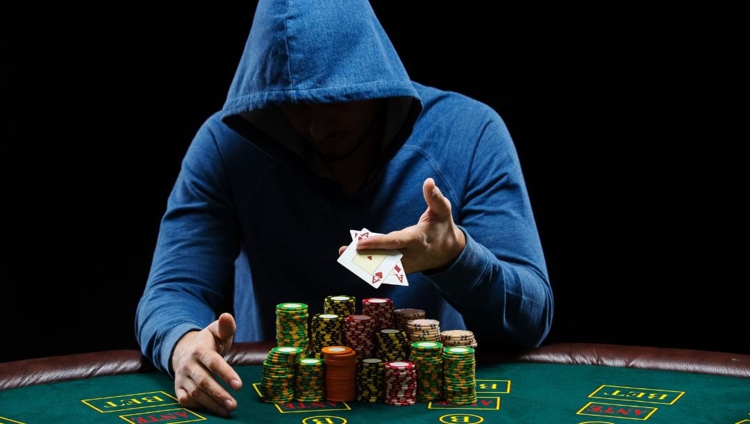 Body image, a man wearing a hoodie that covers his face as he looks down, sitting at a poker table with poker chips stacked in front of him, he holds out a pocket pair of aces 