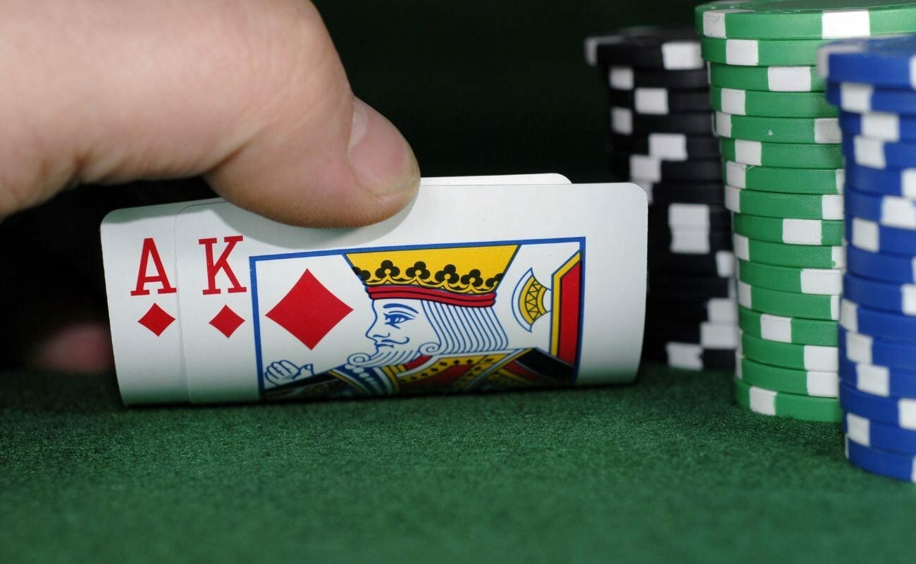 a close up of a man’s finger checking his hole cards, an ace and a king of diamonds, during a game of poker on a green felt poker table next to stacks of poker chips 