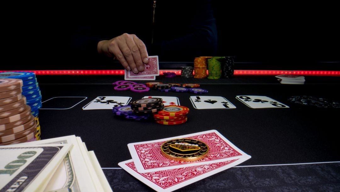 Body image, a poker game taking place from a first person point of view with a man sitting opposite at the poker table, his hands picking up his hole cards, two hole cards and tokens in front of the camera, money and poker chips stacked on the side 